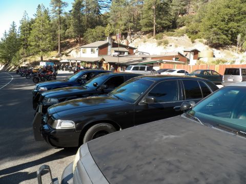 2003 Ford Crown Victoria - Several dark sedans line a gravel lot fronting a rustic mountain roadhouse and motorcycles.
