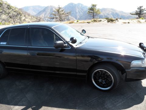 2003 Ford Crown Victoria - Side view of a blacked-out full-size sedan on black wheels parked before rugged mountains.