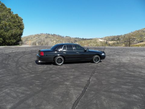 2003 Ford Crown Victoria - Rear-quarter view of a black sedan with antenna mast on an open pad scarred by skid marks.