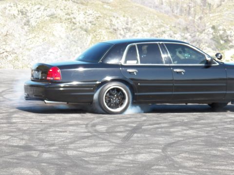 2003 Ford Crown Victoria - Full-size black sedan smokes its rear tire mid-burnout on a hillside asphalt pad.