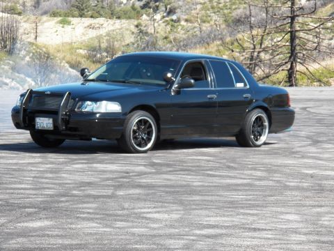 2003 Ford Crown Victoria - Blacked-out interceptor-style sedan with brush guard rests on a tire-marked mountain pad.