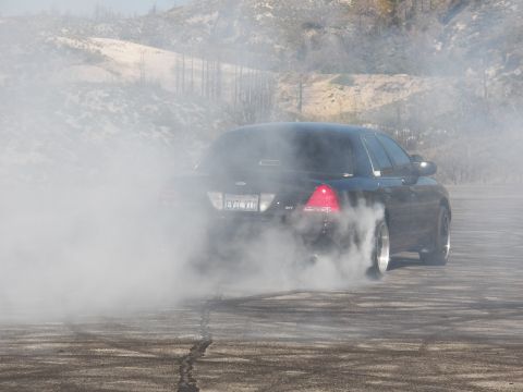 2003 Ford Crown Victoria - Dense white smoke engulfs the rear of a black sedan mid-burnout against dry hills.
