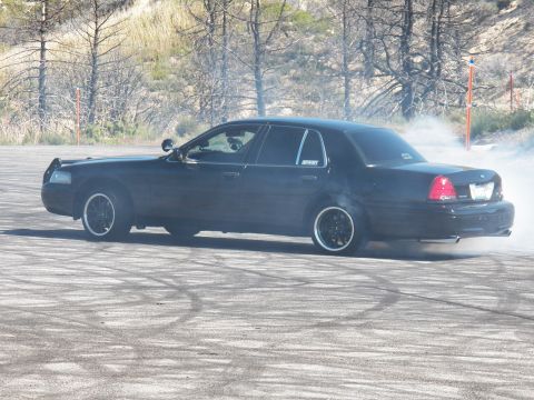 2003 Ford Crown Victoria - Full-size black sedan kicks up smoke through a slide on a hillside burnout lot.