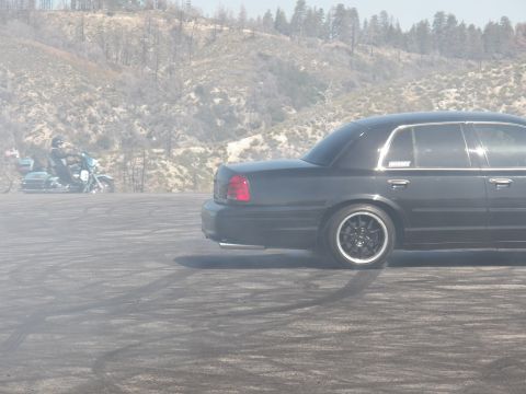 2003 Ford Crown Victoria - Dark full-size sedan smokes a tire while a parked bike sits in the hazy background.