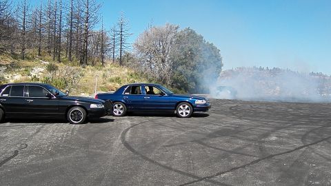 2003 Ford Crown Victoria - Two parked full-size sedans frame a smoking burnout on a tire-streaked mountain pad.