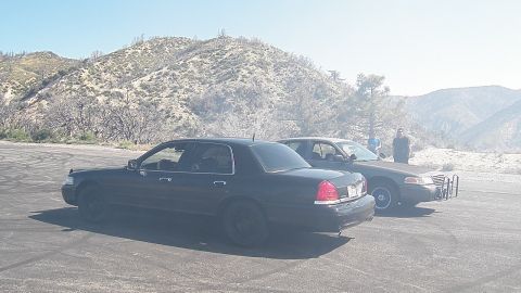 2003 Ford Crown Victoria - Pair of dark full-size sedans staged on an open pad as figures stand in the distance.