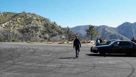 2003 Ford Crown Victoria - Figure strides over an open tire-marked pad toward dark sedans and people at right.
