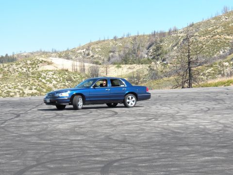 2003 Ford Crown Victoria - Blue four-door sedan slides over an open burnout pad below dry hills, driver visible.