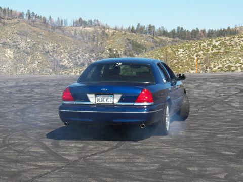2003 Ford Crown Victoria - Blue full-size Ford sedan smokes its rear tire mid-burnout on a skid-marked lot.