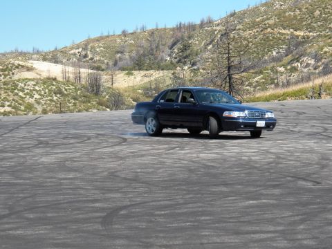 2003 Ford Crown Victoria - Navy full-size Ford sedan carves a slide over an open tire-marked burnout pad.