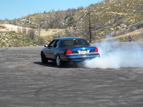 2003 Ford Crown Victoria - Blue four-door kicks up a thick smoke cloud mid-burnout as a bike waits behind.
