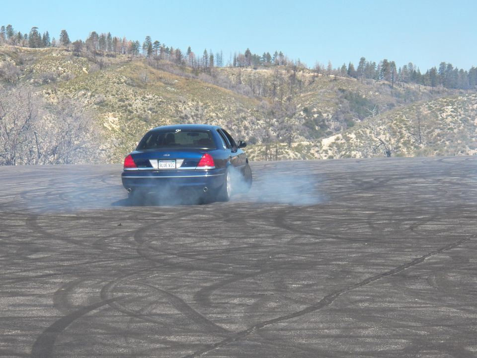 Blue Ford Crown Victoria doing a burnout on tire-marked asphalt lot in hills