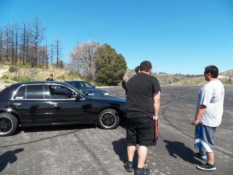 2003 Ford Crown Victoria - People gather near a black full-size sedan parked on a tire-marked asphalt clearing