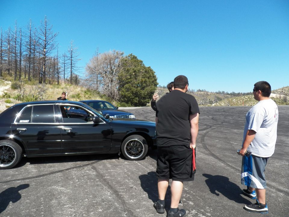 Three men standing beside a black Ford sedan on a mountain lot under blue sky