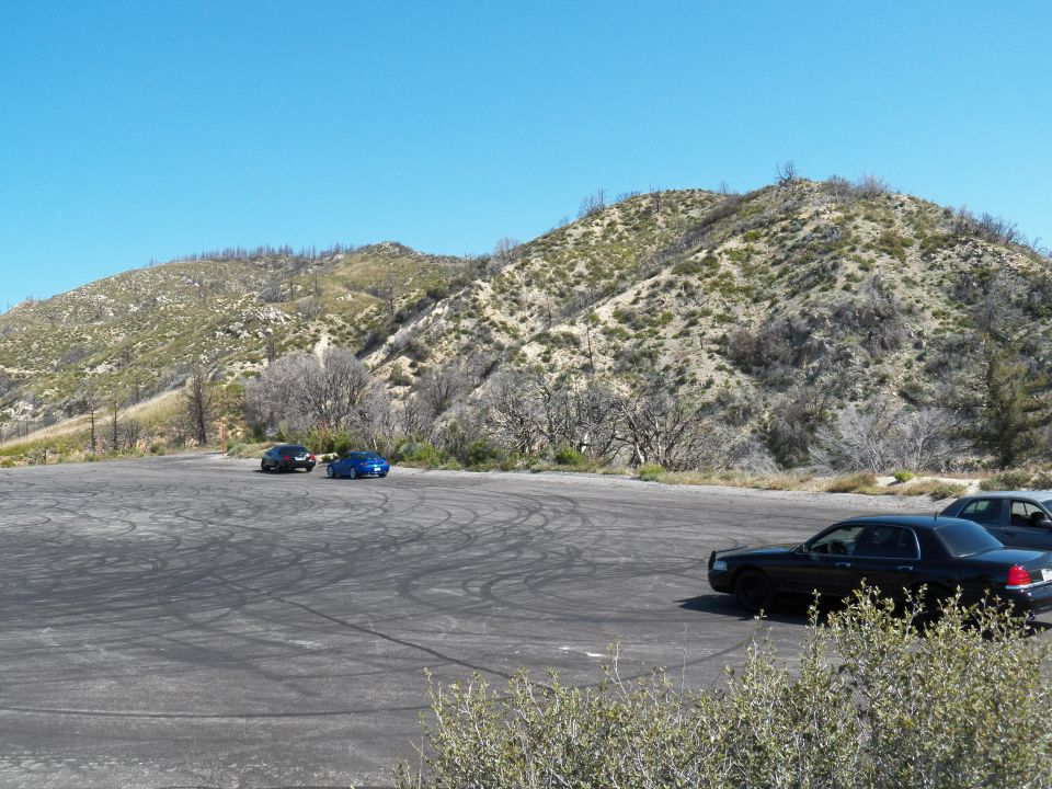 Cars parked across a large tire-marked asphalt lot below brushy hills