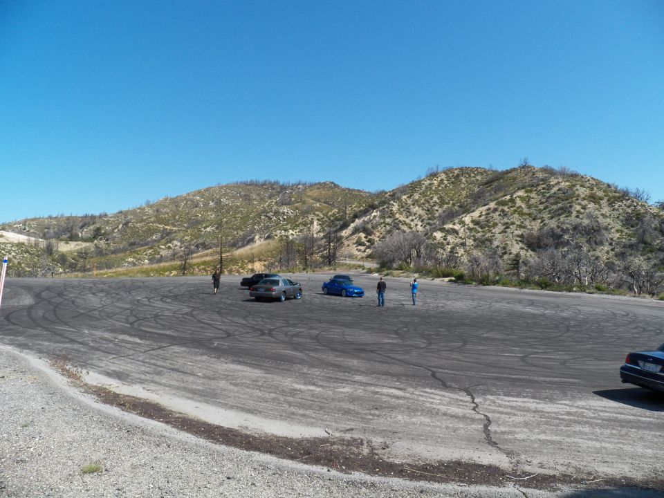 People and cars scattered across a large asphalt lot ringed by brushy hills