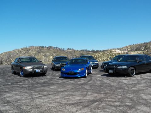 2003 Ford Crown Victoria - Front view of several full-size sedans flanking a blue roadster on asphalt
