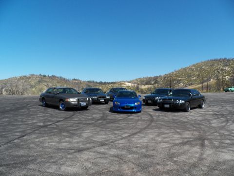 2003 Ford Crown Victoria - Car meetup on a tire-marked dirt lot: dark Crown Victoria-style sedans flank a blue Honda roadster.