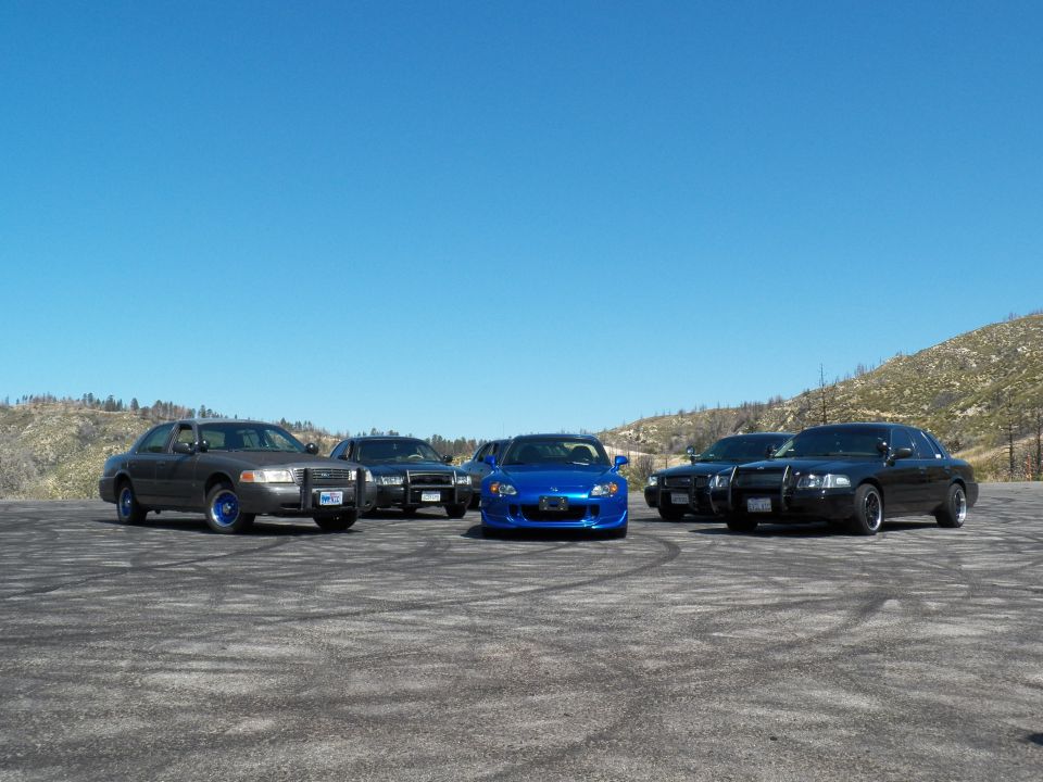 Front lineup of grey and black full-size sedans with a blue Honda roadster center on dirt lot