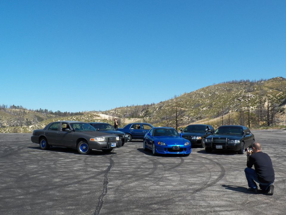 Man kneeling to photograph a blue Honda roadster and dark sedans on a dirt lot