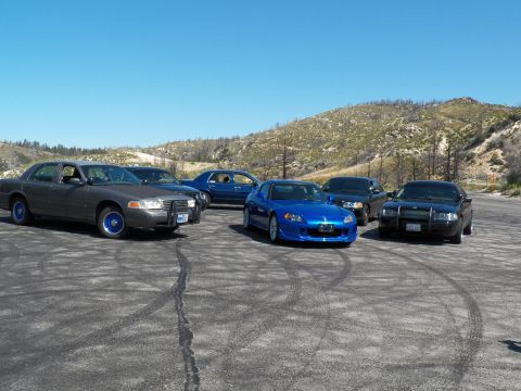 2003 Ford Crown Victoria - Tight grouping of dark full-size sedans around a blue Honda roadster on dirt.