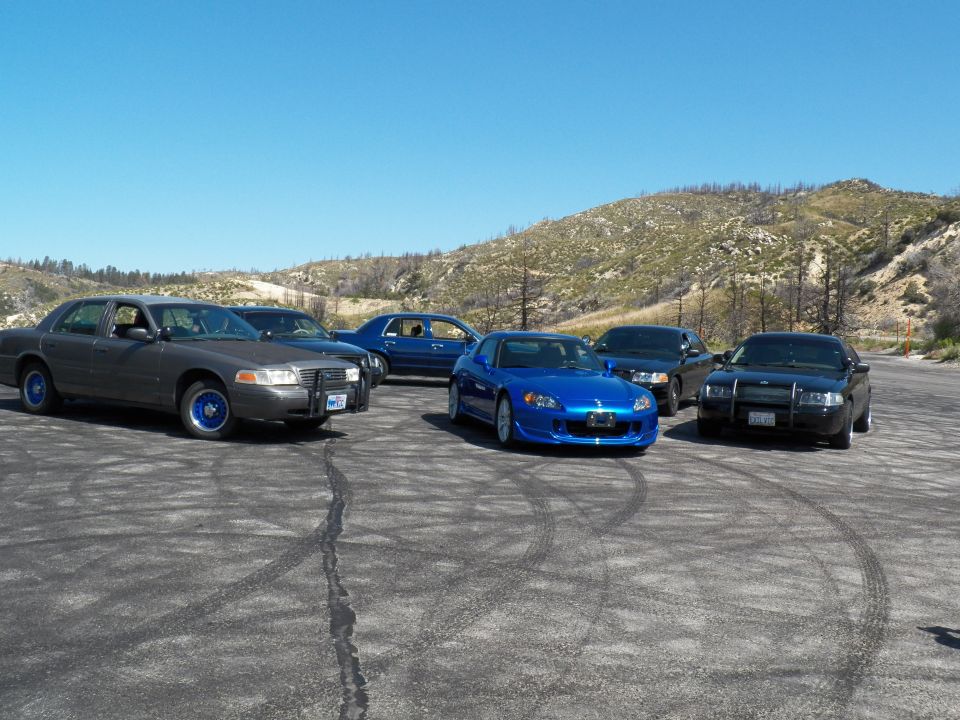 Grey sedan with push bar, blue Honda roadster and black sedans grouped on a gravel lot