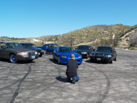 2003 Ford Crown Victoria - Person crouches at a blue roadster ringed by grey and black sedans on gravel.