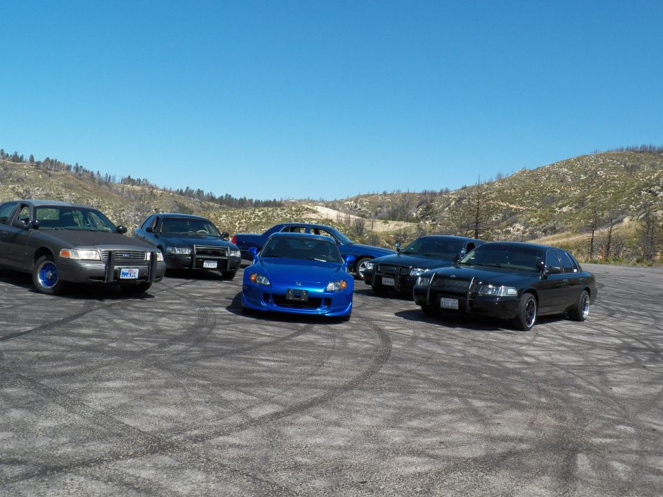 Blue Honda roadster centered among grey and black full-size sedans on a dirt lot