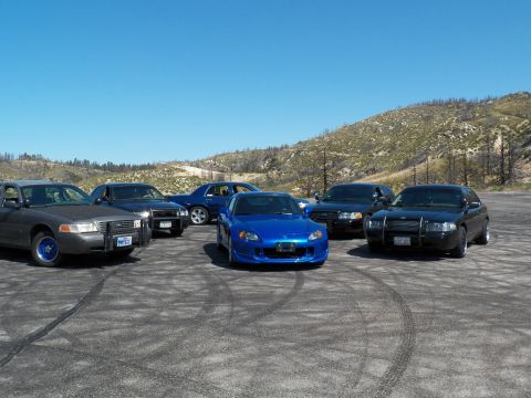 2003 Ford Crown Victoria - Blue roadster faces the camera amid dark sedans with push bars on dirt.