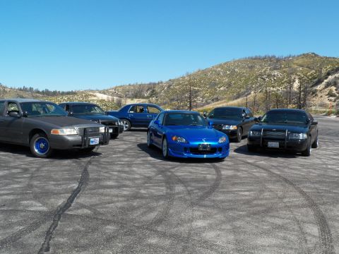 2003 Ford Crown Victoria - Bright blue Honda roadster stands out among darker full-size sedans on dirt.