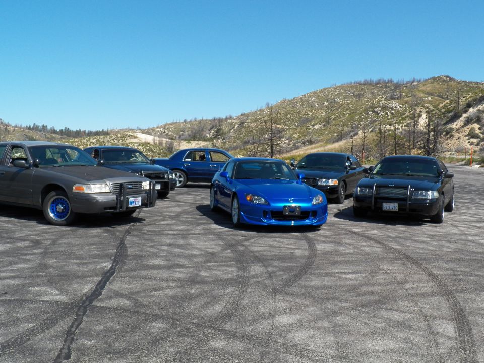 Metallic blue Honda roadster with dark sedans and a blue sedan behind on a gravel lot