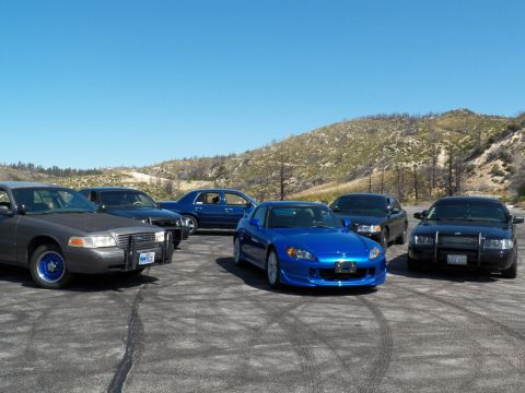 2003 Ford Crown Victoria - Roadster centered between a grey police-style sedan and black sedans on gravel.
