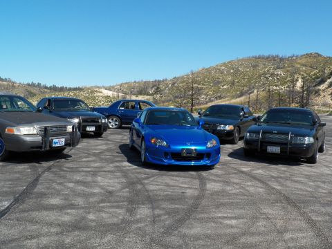2003 Ford Crown Victoria - Front-quarter view of a blue roadster amid grey and black sedans on dirt.