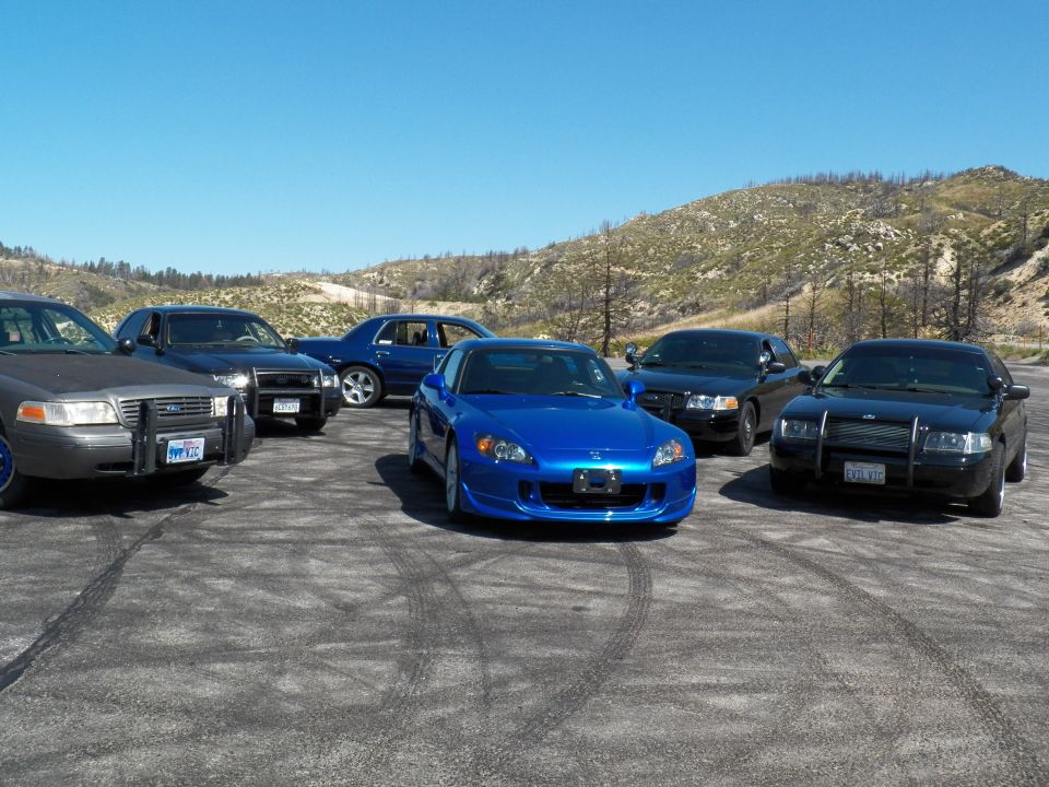 Blue Honda roadster front-three-quarter with dark full-size sedans on a gravel lot