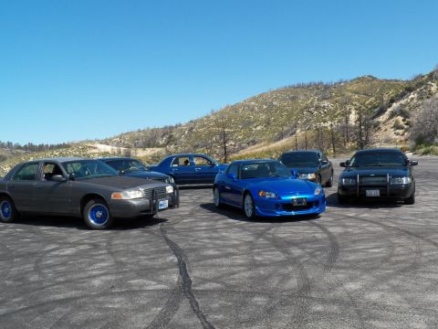 2003 Ford Crown Victoria - Blue roadster and a grey police-style sedan share a gravel lot below dry hills.