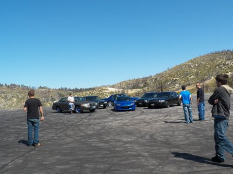2003 Ford Crown Victoria - Onlookers gather near a row of dark sedans and a blue roadster on dirt.
