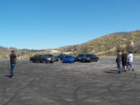 2003 Ford Crown Victoria - Distant lineup of dark sedans and a blue roadster watched by a few bystanders.