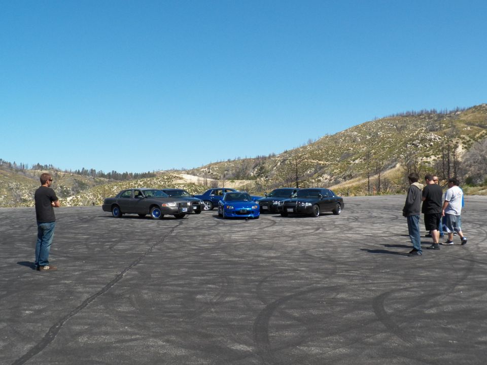 Distant row of dark sedans and a blue roadster with bystanders on a tire-marked gravel lot