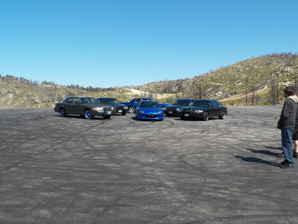 Dark full-size sedans and a blue roadster parked across a wide gravel lot below hills