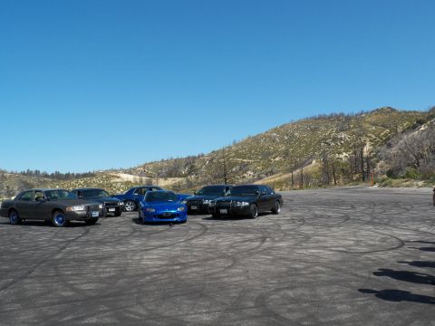 2003 Ford Crown Victoria - Roadster and dark sedans parked at the far edge of a tire-scuffed dirt lot.