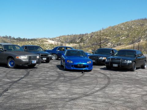 2003 Ford Crown Victoria - Blue roadster faces forward beside a grey push-bar sedan and black sedans.