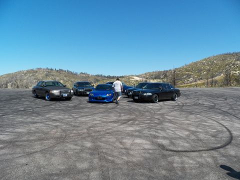 2003 Ford Crown Victoria - person stands beside a blue roadster framed by dark sedans on dirt.