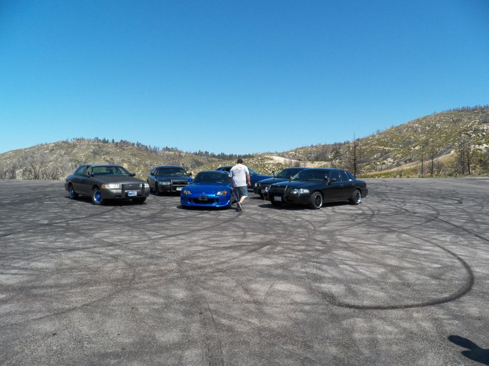 Person standing by a blue roadster among grey and black sedans on a wide gravel lot