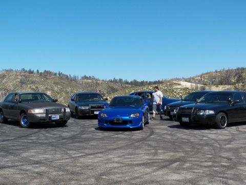 2003 Ford Crown Victoria - Blue roadster anchors a group of dark sedans as a man walks behind on dirt.