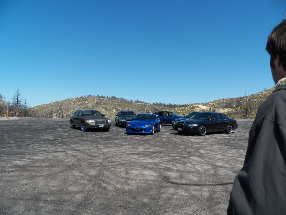 Cars parked in distant gravel lot below dry hills, person in jacket watching at right