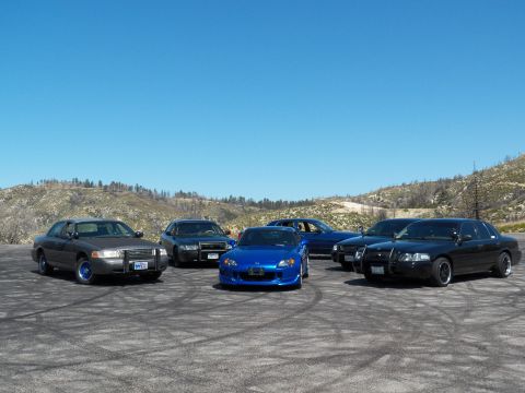 2003 Ford Crown Victoria - Several cars staged on a dirt pullout, bright blue convertible flanked by black sedans
