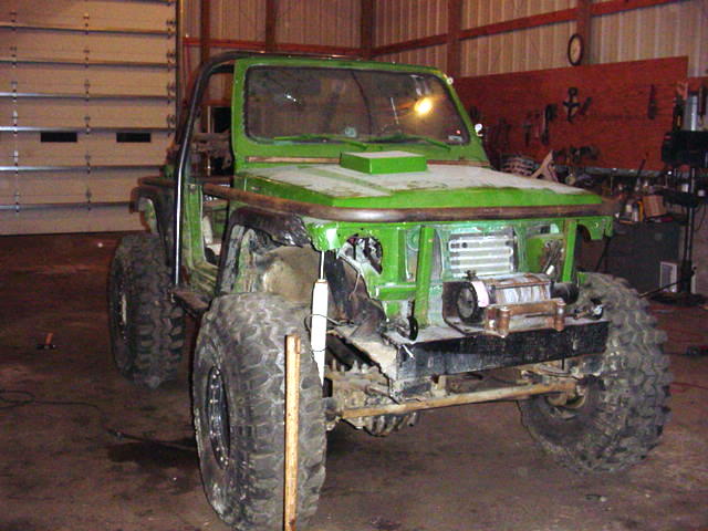 Green off-road vehicle with large tires and roll cage, front end disassembled, in a garage
