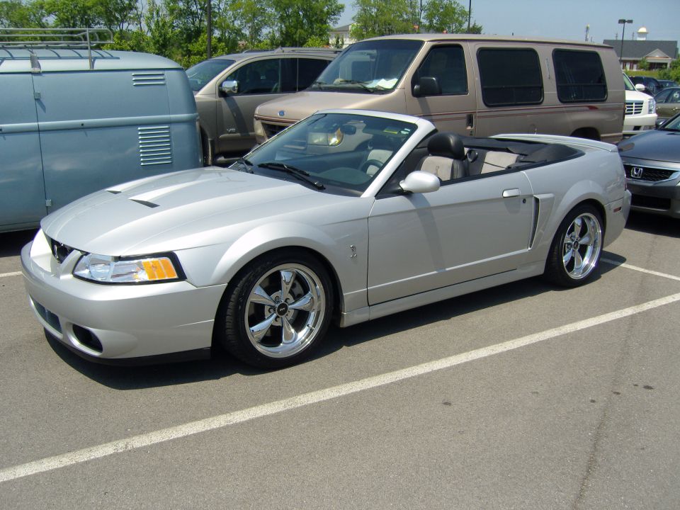 Silver convertible parked in a lot, side view. Other vehicles like a van and a blue camper are visible