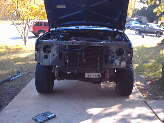 Front of a dark blue Ford truck with hood up, grille and bumper removed. License plate TRW-3430 visible. Red SUV and gray pickup in background