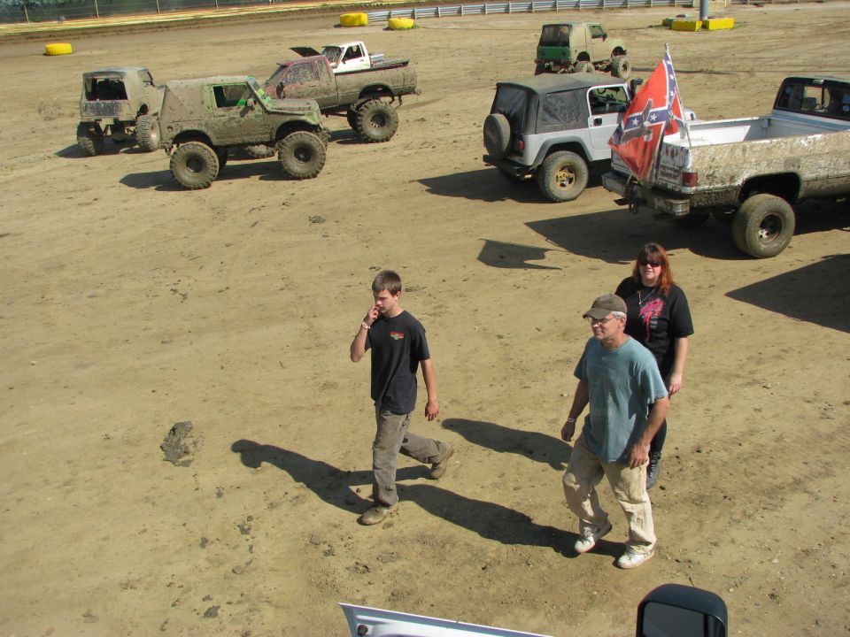 Muddy off-road vehicles parked on dirt, with people walking nearby. A Confederate flag is visible on one truck